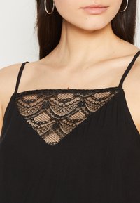 Czarny bezrękawnik z trójkątnym koronkowym wzorem na piersiach, złożonym z misternych motywów. Wykonany z miękkiej tkaniny z regulowanymi ramiączkami.