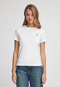 Vit bomull t-shirt med korta ärmar och rund halsringning. Har en liten svart grafisk logotyp på bröstet. Kombinerad med blåjeans.