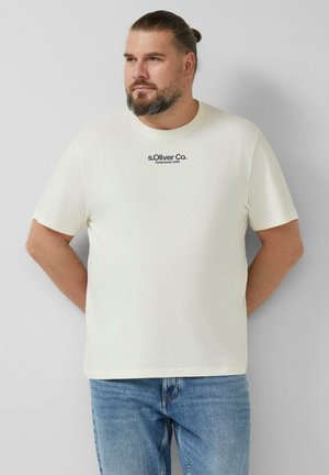 Man with beard and bun wearing cream s.Oliver Co. T-shirt and blue jeans, standing with hands behind back, looking left.