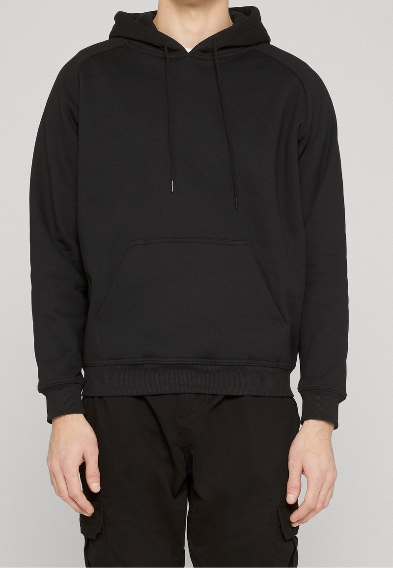 Personne portant un sweat à capuche noir uni avec poche avant et un pantalon cargo noir, sur un fond neutre.
