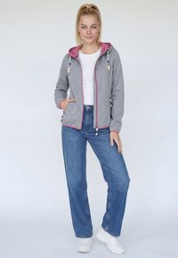 Jeune femme debout, main dans la poche, portant un sweat à capuche gris avec des bordures roses, un t-shirt blanc, un jean bleu et des baskets blanches sur un fond clair.