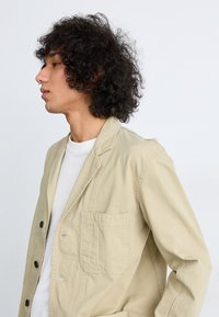 Beige cotton blazer with a single pocket, notch lapels, and five black buttons. Features a relaxed fit and lightweight, textured fabric.