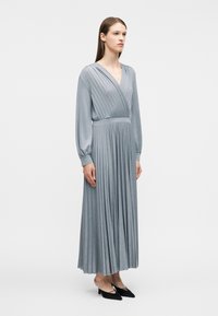 Γυναίκα που στέκεται, φορώντας ένα μακρύ ασημένιο πλισέ φόρεμα με μακριά μανίκια και μαύρα παπούτσια με μυτερή μύτη, σε λευκό φόντο.