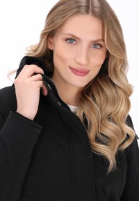 Blond kvinde med bølget hår smiler, mens hun holder kraven på sin sorte jakke mod en simpel hvid baggrund.