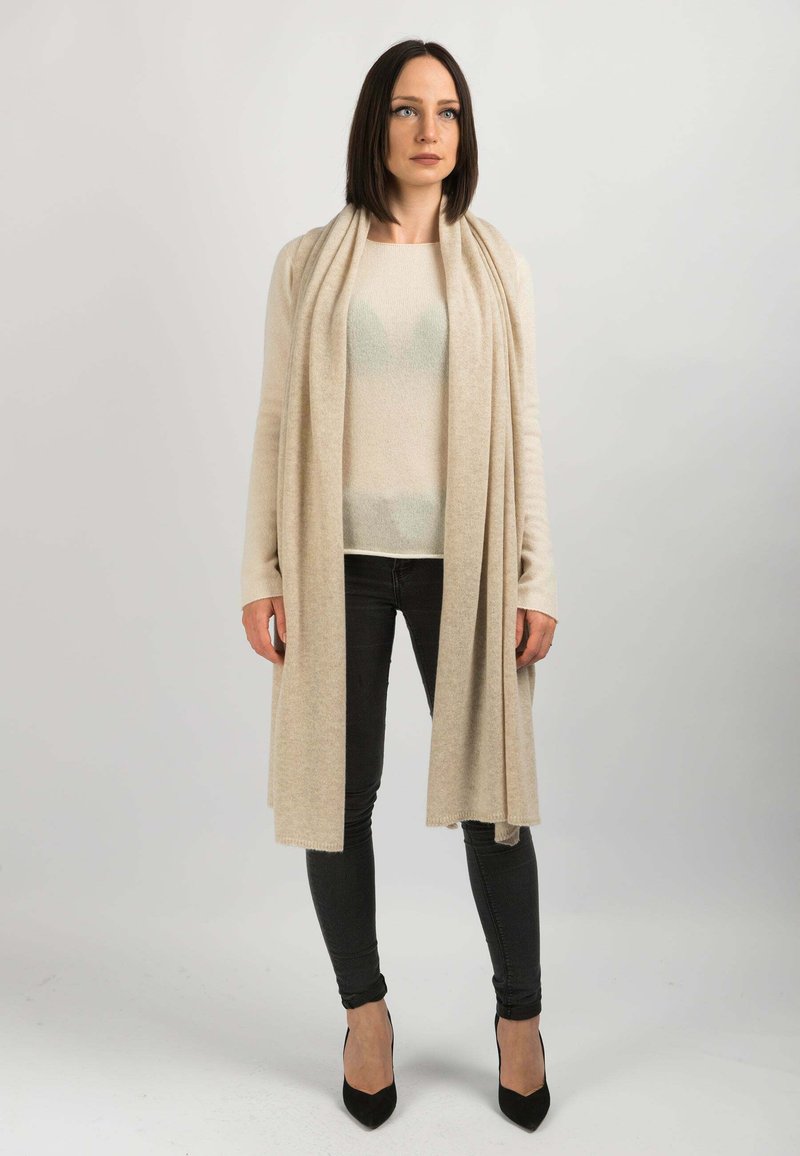 Beige cardigan lavet af blødt stof, der hænger over en lys sweater og sorte jeans, med et langt design og glat tekstur. Sorte hæle.