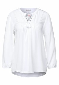 Vit långärmad blus med veck, rund hals, dekorativa spetsdetaljer och fransdetalj vid halsringningen. Mjuk tygtextur.