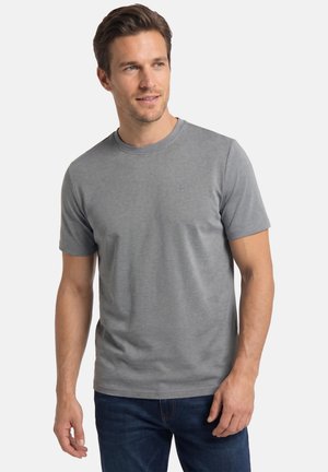Mand med lys hud og kort brunt hår, iført en ensfarvet grå kortærmet skjorte og blå jeans, der kigger lidt til siden.