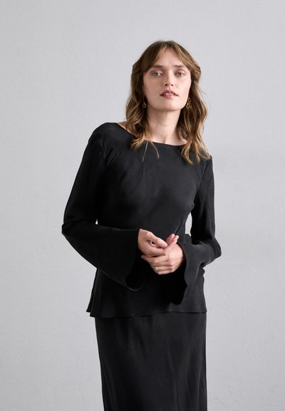 Haut noir à manches longues avec un décolleté bateau et des manches évasées ; design superposé avec une texture lisse. Porté avec une jupe noire.