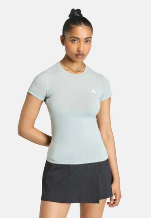 Femme avec les cheveux en chignon portant une chemise ajustée à manches courtes gris clair avec un petit logo et une jupe noire sur un fond uni.