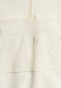 Cremfarbener Sweatshirt mit einer strukturierten oberen Hälfte und einer glatten unteren Hälfte, versehen mit dem weißen, kursiven Schriftzug "Definitely". Mit Reißverschluss am Hals.