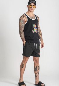 Čierny top bez rukávov s farebnou grafikou medveďa, doplnený čiernymi šortkami so podobným dizajnom medveďa, skombinovaný s šľapkami.