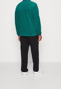 Grön fleecetröja med en mjuk textur, utrustad med rund halsringning och lila detaljer vid ärmsluten. Bärs med svarta byxor och vita sneakers.