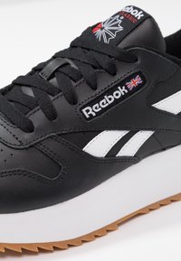 Czarny skórzany sneaker z białymi akcentami i teksturowaną gumową podeszwą. Posiada logo Reebok oraz wyściełany język z czerwono-białym logo.