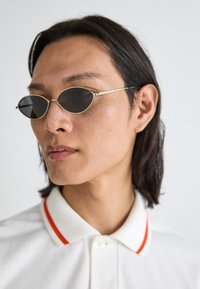 Gafas de sol ovaladas con lentes oscuros, montura de metal plateado, llevadas con una camiseta polo blanca con cuello a rayas rojas. Textura de piel suave.