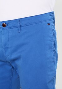 Modré bavlněné šortky s hladkým povrchem, obsahující hnědé zapínání na knoflík, kontrastní stehy a malé logo značky blízko pásku.