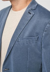 Niebieska marynarka z teksturowanej tkaniny, z klapami notched, pojedynczą kieszenią na piersi i subtelnymi detalami w szwie, w połączeniu z jasnoszaryą koszulą.