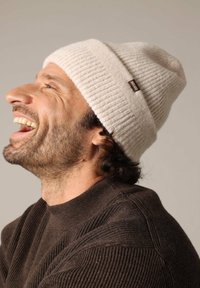 Crème geribbelde beanie met een opgevouwen rand en een klein bruin label. Gemaakt in combinatie met een bruine gestructureerde trui, die een casual, relaxte pasvorm toont.