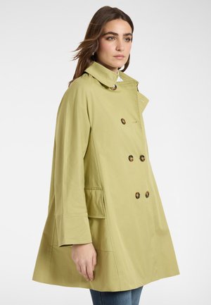 Mujer con cabello castaño lleva un abrigo trench doble botonadura de color verde claro con botones grandes, de pie contra un fondo blanco.