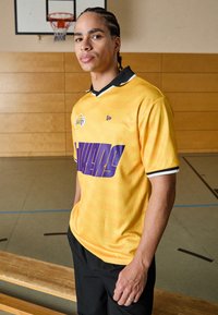 Gelbes Basketball-Trikot mit lila "LAKERS"-Schriftzug, schwarzem Kragen mit weißem Saum und Logodetail. Tragen mit schwarzen Hosen, in einem Sportgymnasium.