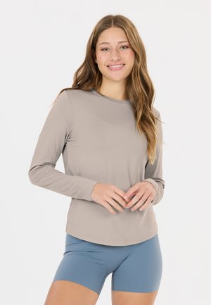 Jeune femme aux longs cheveux sourit, portant une chemise beige à manches longues et un short de sport bleu sur un fond clair uni.