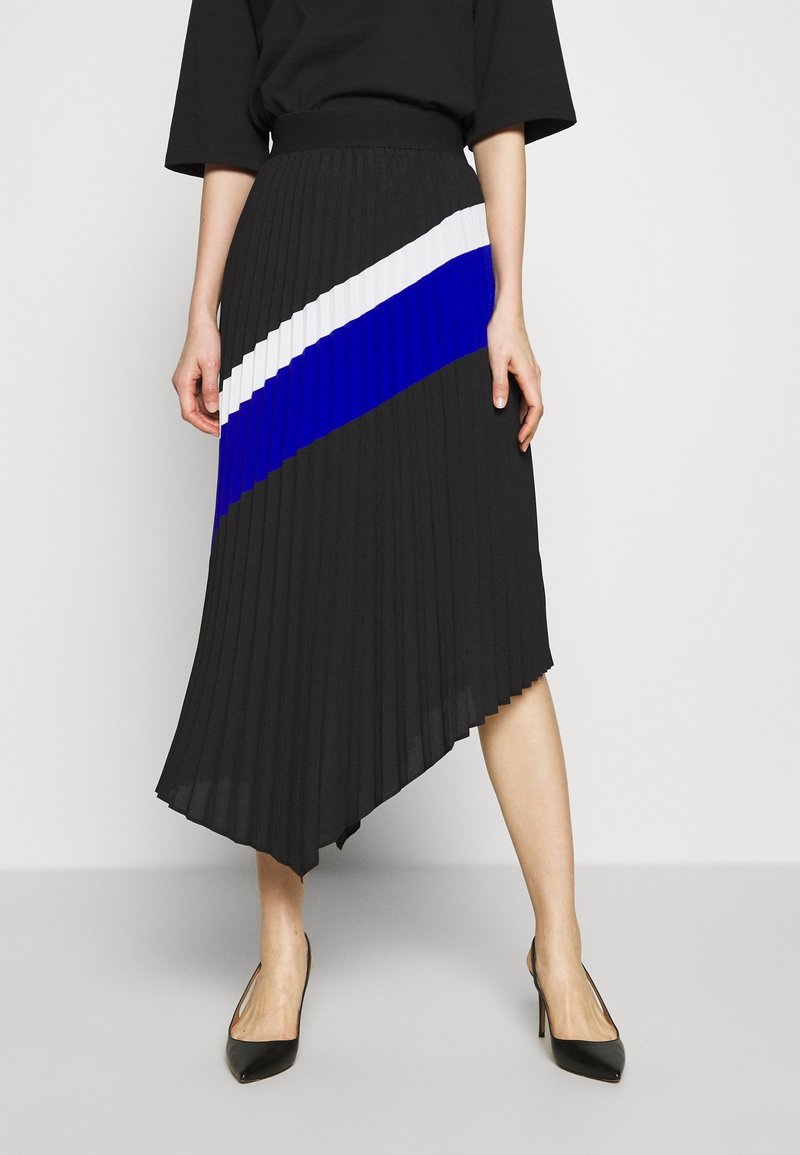 Femme portant une jupe asymétrique plissée noire avec des rayures diagonales blanches et bleues, associée à des talons hauts noirs pointus.