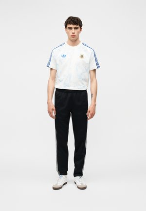 Jeune homme debout face à l'avant, portant un t-shirt blanc Adidas avec des bandes bleues, un pantalon de survêtement noir Adidas avec des bandes blanches et des baskets blanches.