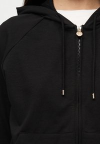 Μαύρο φούτερ με φερμουάρ, κουκούλα με κορδόνι, χρυσά εξαρτήματα φερμουάρ και πλεκτή υφή. Διαθέτει ραγκλάν μανίκια και μπροστινή τσέπη.
