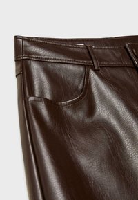 Gros plan sur un pantalon en cuir marron foncé montrant la poche avant, la ceinture avec passants de ceinture et les coutures détaillées sur une texture en cuir lisse.