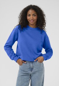 Femme avec des cheveux bouclés portant un sweat-shirt bleu vif et un jean bleu clair, les mains dans les poches, souriante devant un fond uni.