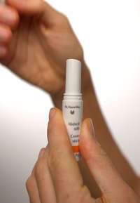 Dr. Hauschka cover stick in a white and orange tube. Compact design with text on the label. Held between fingers against a blurred background.