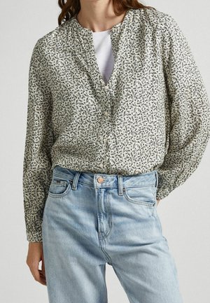 Frau trägt eine helle, geblümte Bluse mit Knöpfen über einem weißen Oberteil, kombiniert mit hoch taillierten, hellblauen Jeans aus Denim.