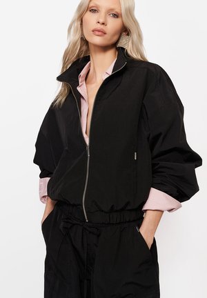 Blondynka w dużej czarnej kurtce na zamek nałożonej na różową koszulę w paski, z rękami w kieszeniach i dużymi kwadratowymi kolczykami.