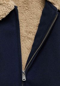 Granatowa kurtka z beżowym, puszystym wyściółką Sherpa, z metalowym zamkiem błyskawicznym i kołnierzem. Teksturowany materiał i kontrastujące kolory podkreślają detale.