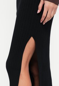 Czarna, prążkowana dzianinowa sukienka z wysokim rozcięciem po boku, na zdjęciu dłoń opiera się na materiale, podkreślając fakturę i detale wzoru.