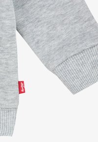 Tessuto del maglione grigio con una finitura texturizzata. Polsini e orlo a coste. Presenta una piccola etichetta rossa sul lato. Design semplice senza motivi.