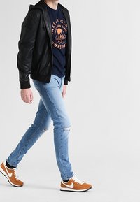 Giacca bomber in pelle nera, t-shirt grafica blu navy, jeans strappati azzurri chiari e sneakers Nike arancioni con dettagli bianchi, che cammina su uno sfondo neutro.