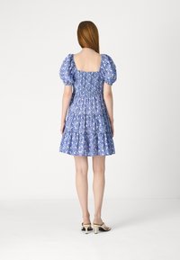 Μπλε floral φόρεμα με φουσκωτά μανίκια, σφηκωτό πίσω μέρος και tiered φούστα. Φτιαγμένο από ελαφρύ ύφασμα με σχέδιο. Ορατά ψηλά παπούτσια.