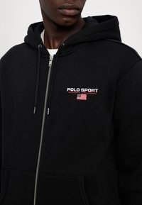 Černá mikina na zip z bavlny, s předním kapsou, nastavitelnými stahovacími šňůrkami a vyšitým logem „POLO SPORT“ s detailním motivem vlajky.