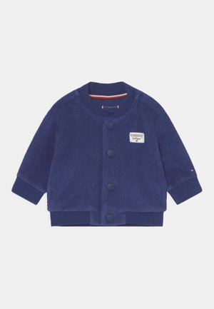 Chaqueta de forro polar azul marino con cuello redondo, cierre de botones y un pequeño parche con el logo de Tommy Hilfiger. Presenta puños y dobladillo acanalados.