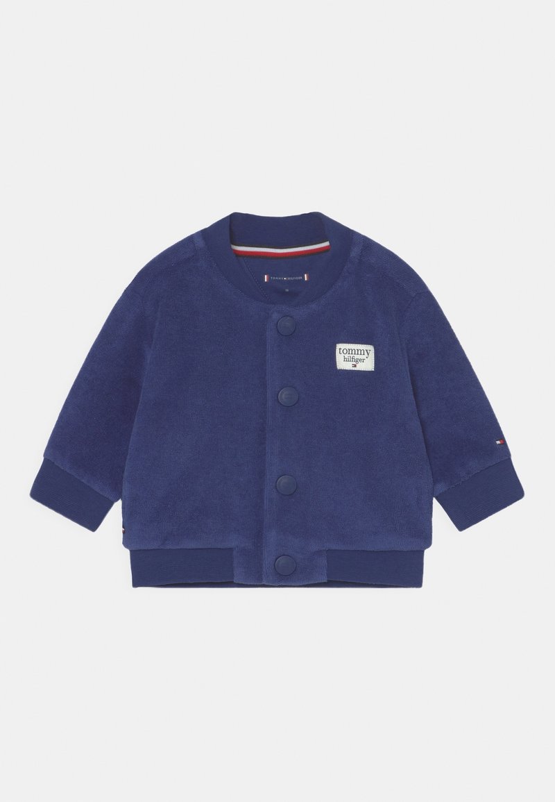 Granatowa polarowa kurtka z okrągłym dekoltem, zapięciem na guziki i małą naszywką z logo Tommy Hilfiger. Posiada ściągacze przy mankietach i u dołu.