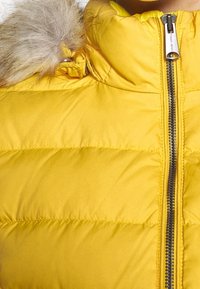 Żółta pikowana kurtka z kapturem wykończonym futerkiem i metalowym zamkiem błyskawicznym z napisem "Tommy Jeans" zamkniętym z przodu.