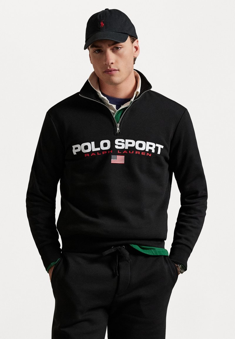 Juodas zip-up džemperis su „POLO SPORT“ didelėmis balta ir raudona spalvomis. Suderintas su juodomis kelnėmis ir žalia palaidine.