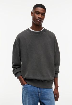 Młody mężczyzna w ciemnoszarym swetrze z okrągłym dekoltem, białej koszuli i niebieskich dżinsach, stojący z jedną ręką w kieszeni na jednolitym białym tle.