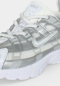 Άσπρο και ασημί αθλητικό παπούτσι Nike με ύφασμα διχτυώματος και συνθετικές επικαλύψεις, υφασμένο πέλμα και λευκά κορδόνια δεμένα πάνω.