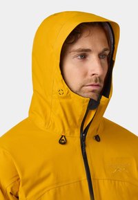 Giacca impermeabile gialla con cappuccio regolabile, dotata di accessori neri, chiusura con zip e dettaglio del logo sul petto. Tessuto liscio e leggero.