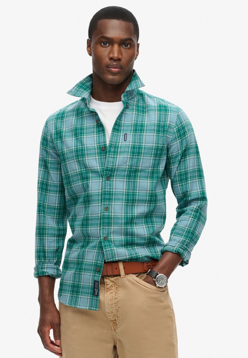 Camicia a quadri verde e blu con bottoni in tessuto leggero, caratterizzata da un colletto piegato, taschino sul petto, abbinata a pantaloni beige e una cintura marrone.