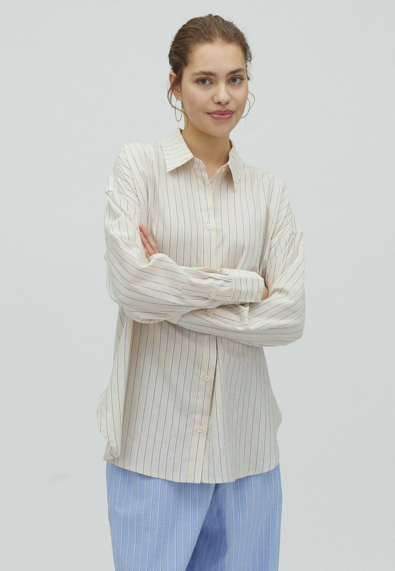Jeune femme aux cheveux châtain clair en chignon, portant une chemise beige rayée et un pantalon rayé bleu, les bras croisés, souriant légèrement, sur fond blanc.