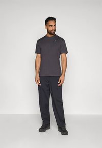 Muž stojaci proti bielej stene, oblečený v tmavosivom tričku s krátkym rukávom, čiernych cargo nohaviciach a čiernych turistických topánkach, s pohľadom do strany.