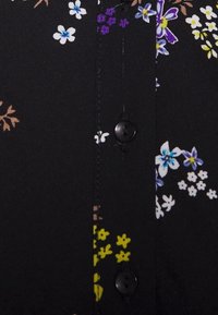 Černá látka se květinovým vzorem v barvách bílé, fialové a žluté. Detail knoflíku viditelný podél švu.