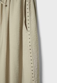 Pantalon en tissu beige texturé avec une poche latérale et des clous métalliques argentés décoratifs courant verticalement le long de la jambe extérieure.